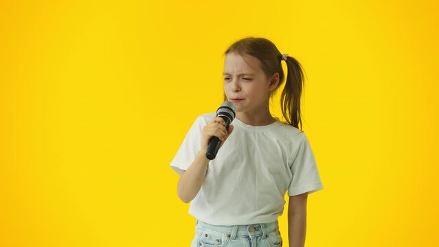 Young little girl passionately singing a song to the microphone and smiling on a yellow background. Talented child showing her creative skills