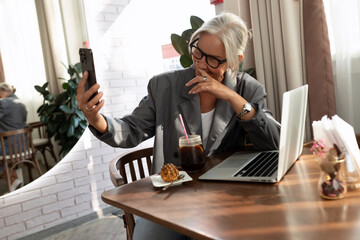 charming stylish mature business woman with glasses dressed in a gray jacket spends time in a cafe sitting with a laptop