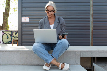 successful european gray-haired senior business woman with a stylish hairstyle and glasses is dressed in a gray jacket sits with a laptop on the street