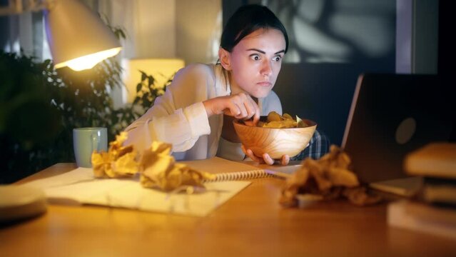 Addicted focused young woman watching interesting program film movie on laptop computer and eating chips alone at home Enchanted female cannot take eyes off screen with snack at late night indoors