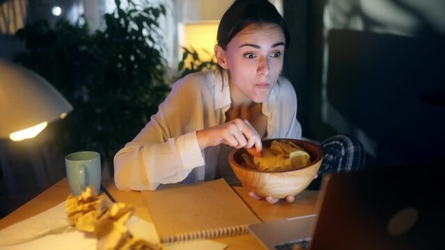 Happy positive woman with chips laughing while watching comedy movie show serials instead of work or education at home workplace Tired female relaxed after hard work day with snack at night indoors