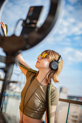 Teenage girl with red glasses, social media blogger recording video, dancing and speaking looking at smartphone on tripod with ring light, on the balcony of the building.