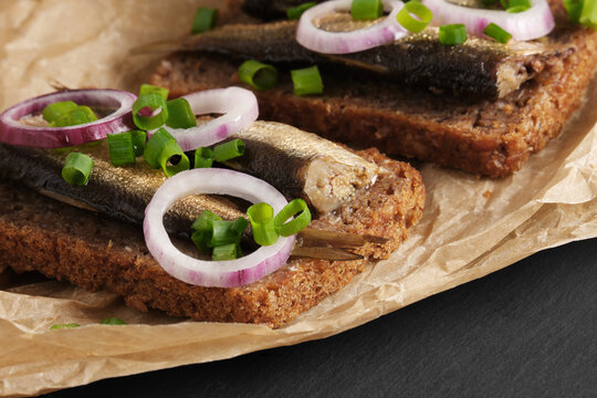 Sandwiches with sprats, red and green onions on baking paper on black stone background. Appetizer, seafood, front view
