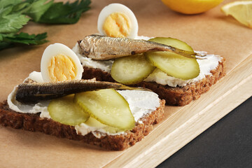 Sandwiches with sprats, quail eggs, pickles on wooden serving tray on black stone background.