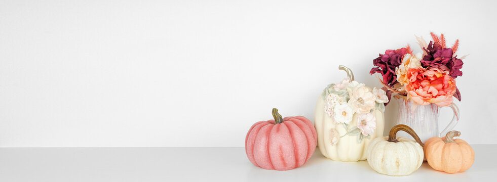 Autumn decor on a white shelf against a white wall banner background. Pumpkins and flowers in vases with pink hue fall colors. Copy space.