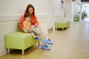 A patient and her baby sit in the clinic corridor, watching videos on their phone while waiting for...