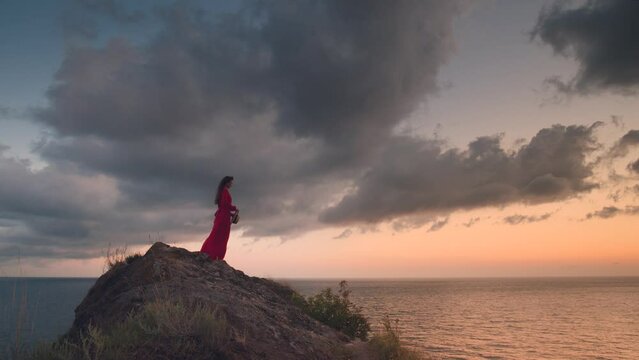 Happy romantic woman standing on the upper coast, sunset footage. The sea wind develops the dress and hair of a girl who mysteriously looks into the distance