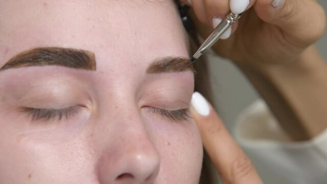A cosmetologist paints a woman's eyebrows in a cosmetology clinic, close-up view. Apply black paint to the eyebrows along the white outline using a brush. Permanent makeup