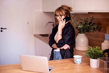 Blond haired mid aged woman standing in the kitchen at home and making a call