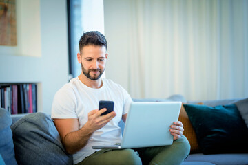 Handsome man sitting at home and using a laptop and mobile phone for work