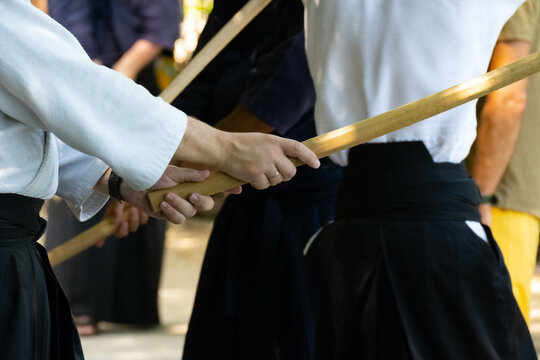 in the rays of the bright sun, the hands of a man in a white uvagi jacket and a black hakama grip a wooden sword during aikido training against the background of other people, the face is not visible