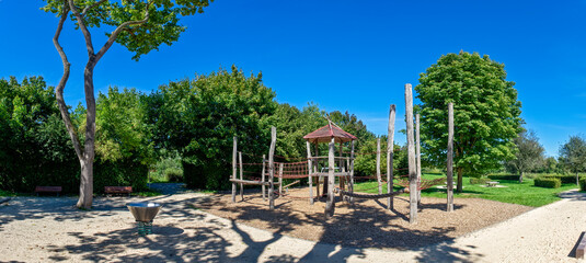 Kletterburg aus Holz auf einem menschenleeren, &ouml;ffentlichen Spielplatz im Sommer bei sch&ouml;nem Wetter und wolkenlosem Himmel, einges&auml;umt von Laubb&auml;umen