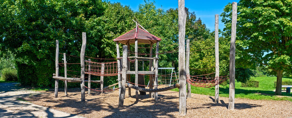 Kletterburg aus Holz auf einem menschenleeren, öffentlichen Spielplatz im Sommer bei schönem Wetter und wolkenlosem Himmel, eingesäumt von Laubbäumen