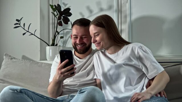 Happy Caucasian married pair sit on couch and remember pleasant moments together.  Handsome man and attractive woman looking at photos in gallery or in social media on gadget and smiling.