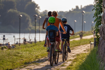 A happy family rides bicycles along the river bank on a summer day towards the sun. Lifestyle and travel concept. Rear view of a group of bikers of different ages wearing helmets. Close-up