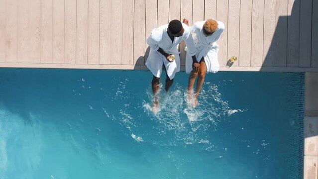 Aerial drone shot of couple wearing robes outdoors sitting with drinks by swimming pool on spa day - shot in slow motion