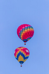 2 hot air balloons are rising in the early morning light into a blue and wispy clouded sky, 1 balloon is above the balloons, The are brightly colored.
