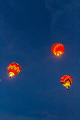 Fototapeta premium 3 hot air balloons are rising in the early morning light into a dark blue and wispy clouded sky. The 3 balloons are brightly lit with their gas burners on, The are brightly colored.