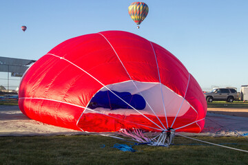 Obraz premium A red hot air balloon is on the ground being deflated. Ropes are coming from the entrance of the balloon. The interior is white and blue. 2 hot air balloons are in the background.