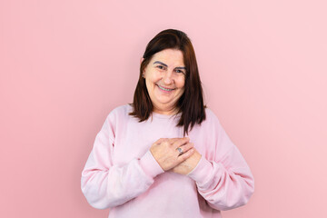 A mature woman, radiating joy and strength, smiles with her hands on her chest against a pink background. An emblem of hope and support for breast cancer awareness and October pink campaigns