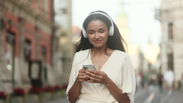 Portrait of young woman with headphones listening to music and hold smartphone scrolling social media online at urban city Pretty female relax enjoying great day and looking around at street outdoors