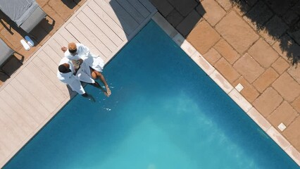Aerial drone shot of couple wearing robes outdoors sitting with drinks by swimming pool on spa day - shot in slow motion - Powered by Adobe
