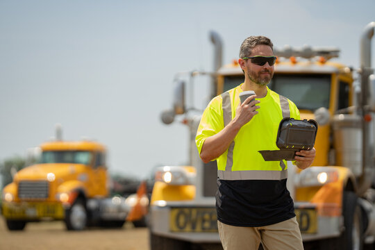 Men driver near lorry trucks. Man owner truck driver in safety vest satisfied near truck. Man driver with lunch box. Truck driver having take away lunch drink coffee to go. Man in front of truck.