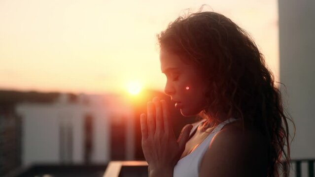 Portrait of hopeful young curly woman clasping hands in prayer asking for blessing and help while the rays of rising sun fall on her face at balcony outdoors Religion and faith concept