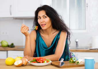 Obraz na płótnie Canvas Portrait of positive asian woman in nightie eating salad in kitchen at home. Woman has cooked fresh vegetable salad.