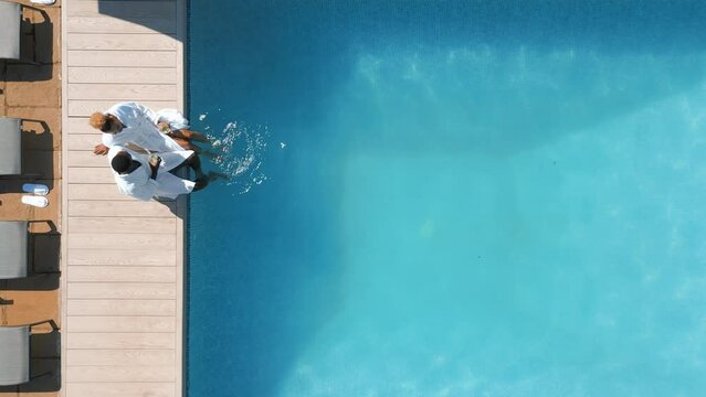 Aerial drone shot of couple wearing robes outdoors sitting with drinks by swimming pool on spa day - shot in slow motion