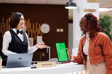 African american woman using tablet with greenscreen display, showing blank copyspace at reception desk with hotel staff. Young female guest holding device with chromakey template.