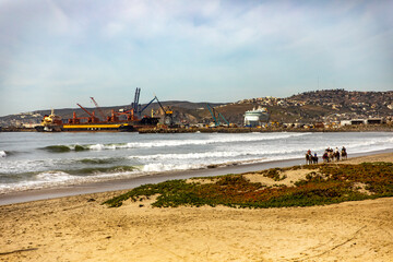 A beautiful beach in Ensenada with the port in the background and people riding horses by the sea, this is one of the best tourist places in Baja California, Mexico.