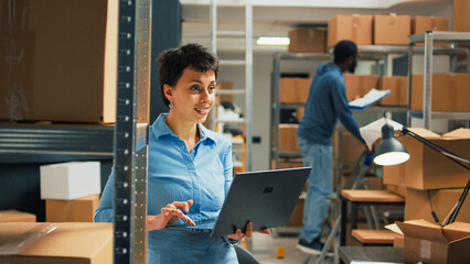 Warehouse owner checking cardboard boxes from storage space shelves, using laptop to plan stock...