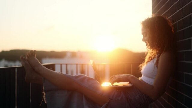 Pretty young woman freelancer working online typing browsing on laptop with beautiful sunset or sunrise at balcony Positive female enjoying great working day at cozy home