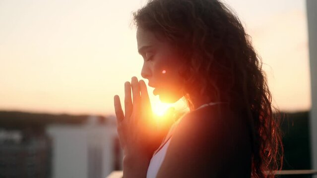 Portrait of hopeful young curly woman clasping hands in prayer asking for blessing and help while the rays of rising sun fall on her face at balcony outdoors Religion and faith concept