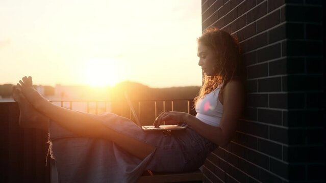 Pretty young woman freelancer working online typing browsing on laptop with beautiful sunset or sunrise at balcony Positive female enjoying great working day at cozy home