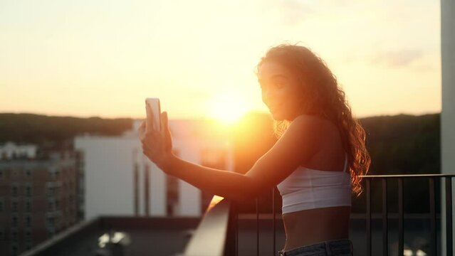 Positive curly female talk on smartphone by video call conference with family or friends with beautiful sunset or sunrise at home balcony Pretty woman blogger create content for social media outdoors
