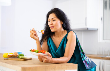 Obraz na płótnie Canvas Asian woman leaning on kitchen table and eating salad. Woman eating vegetable salad in morning, healthy breakfast.