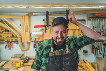 Smiling male carpenter with a clamp on his head, workshop background. High quality photo