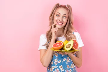 Fotobehang Photo of minded nice woman dressed print overall hold plate with fruits look empty space finger teeth isolated on pink color background © deagreez