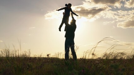 Fototapeta na wymiar Silhouette, father child son, play together in park in front of sun. Dad plays with his son, throws child up into sky with his hands, happy child smiles. Concept of family happiness. Child is flying