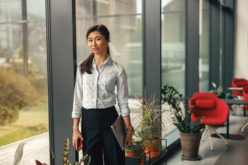 Portrait of stylish business woman standing with laptop near office window. High quality photo