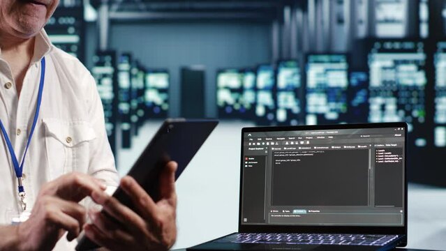 Senior technician in data center, solving tasks on tablet while running scripts on laptop terminal. Licensed serviceman amidst servers, accessing databases and manipulating lines of code