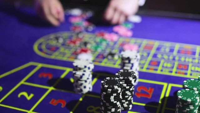 Vibrant casino table with roulette in motion, with casino chips, tokens, the hand of croupier, dollar bill money and a group of gambling rich wealthy people playing bet in the background
