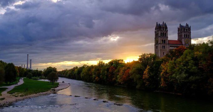 Timelapse of Munich at dramatic sunset: autumn in famous tourist landmark Bavarian city. View of Isar river, trees, and church from Reichenbach Bridge. Munchen, Bavaria, Germany. Camera zoom in