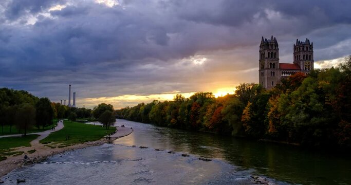 Timelapse of Munich at dramatic sunset: autumn in famous tourist landmark Bavarian city. View of Isar river, trees, and church from Reichenbach Bridge. Munchen, Bavaria, Germany, Europe