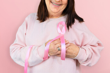 Mature woman smiles, holding a pink ribbon symbolic of breast cancer prevention, emphasizing Pink October awareness.She is isolated against a pink background.