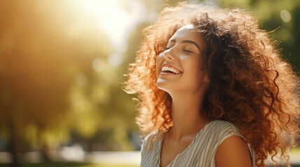 On a sunny day, a young woman with curly hair is standing outside in a park and laughing.