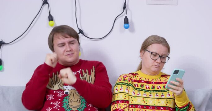 Married couple in ugly sweaters sit on sofa in quarrel, relationship difficulties, differences in temperament. Simpleton man funny flirting with introvert with smartphone Husband amuses offended wife