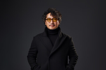 Portrait of a smiling Asian businessman wearing glasses and black suit with looking at the camera in confidently while standing alone in a dark wall background.